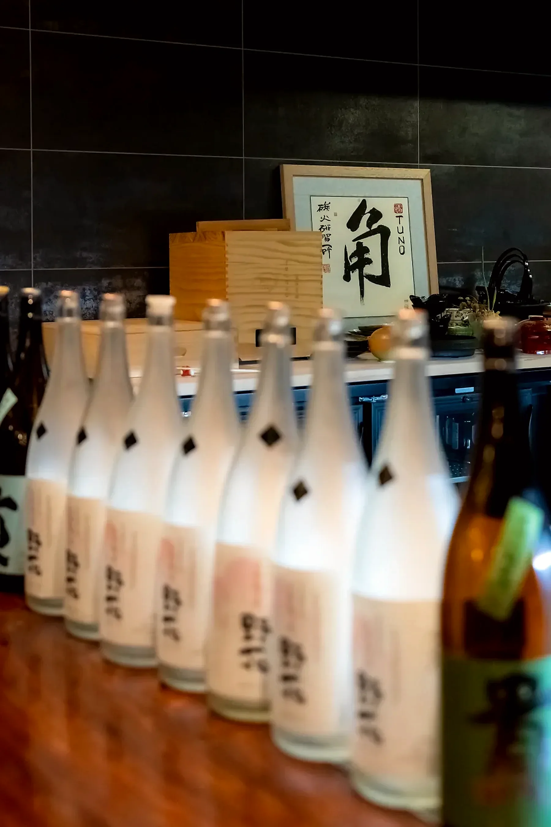 Sake bottles along the bar in front of the framed TUNO calligraphy