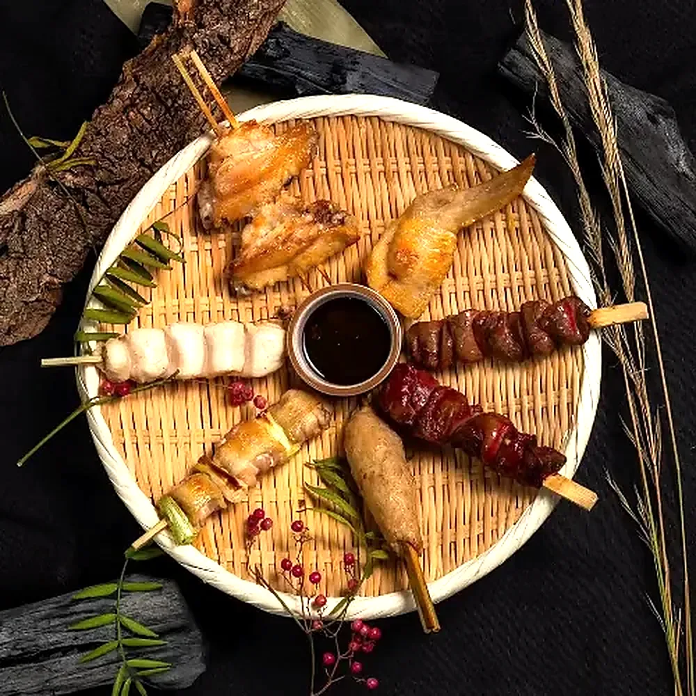 A bamboo basket of grilled skewers with dipping sauce, charcoal lying alongside